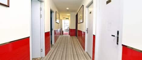 A hotel corridor with doors on both sides, a carpeted floor, and a view into a room with a bed and a window.