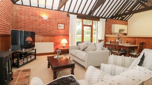 A cozy living room with a brick wall, wooden beams, a television, a sofa, a coffee table, and a dining area with a table and chairs. - Pear Tree (Dereham)