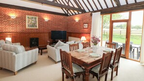 A cozy room with a brick wall, wooden ceiling beams, a television, sofas, a dining table set for four, and a view of the outdoors. - Pear Tree (Dereham)
