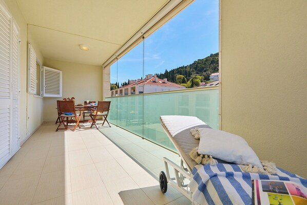 A balcony with a glass railing, a small table with chairs, and a bed with a striped blanket and pillows. - Hedera Estate, Hedera A58 (Dubrovnik)