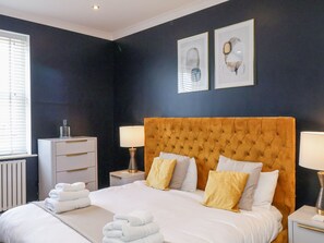 Una cama bien hecha con sábanas blancas y varias almohadas, una mesita de noche con lámpara, un aparador blanco y una obra de arte en la pared.