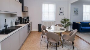 A modern kitchen with a dining area, featuring a round table with chairs, a coffee machine, and a radiator. - The Salisbury - 1 Bedroom Deluxe Apartment (Scarborough)