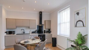 A modern kitchen with a central island, a dining area with a round table and chairs, and a large window with blinds. - The Salisbury - 1 Bedroom Deluxe Apartment (Scarborough)