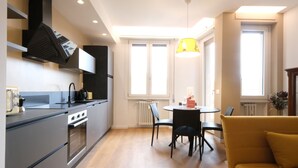 Een moderne keuken met een ingebouwde oven en kookplaat, een zithoek met een ronde tafel en stoelen, en een gele bank.
