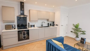 A modern kitchen with a built-in oven, microwave, and coffee maker. There is a blue sofa and a small table with a plant. - The Salisbury 1 Bed Classic Apartment (Scarborough)
