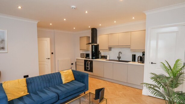 A modern living room with a blue sofa, a glass coffee table, and a kitchen area with white cabinets and a black range hood. - The Salisbury 1 Bed Classic Apartment (Scarborough)