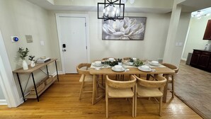 Un coin repas avec une table en bois dressée pour quatre personnes, entourée de chaises en bois. On y trouve une porte blanche avec une serrure, une petite table d’appoint avec des plantes et un tableau accroché au mur.