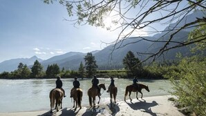 A group of people riding horses along a riverbank with mountains in the background. - Boutique Hotel Engadinerhof – A Home for Generation (Pontresina)