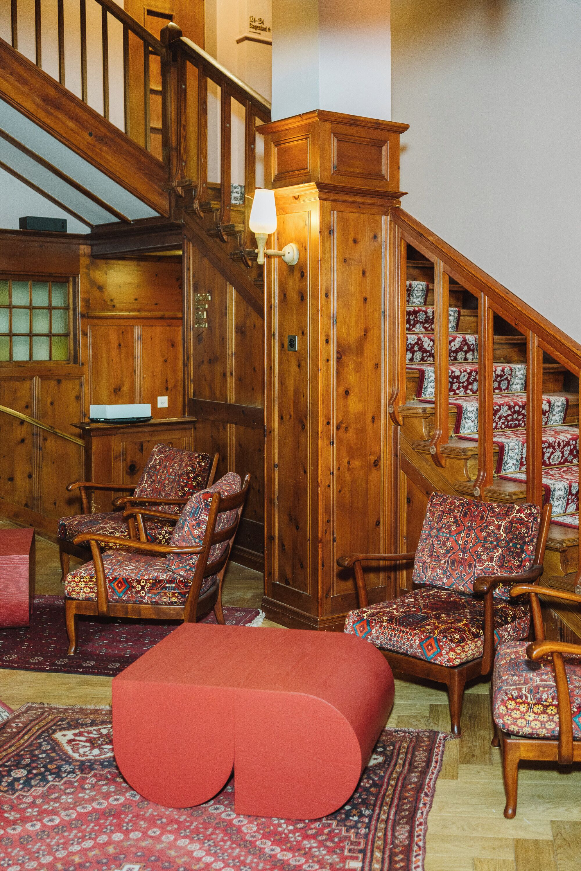 Una escalera de madera con escalones alfombrados y un estampado, una silla de madera con cojín floral y un reposapiés rojo en una habitación con paredes y suelo de madera.