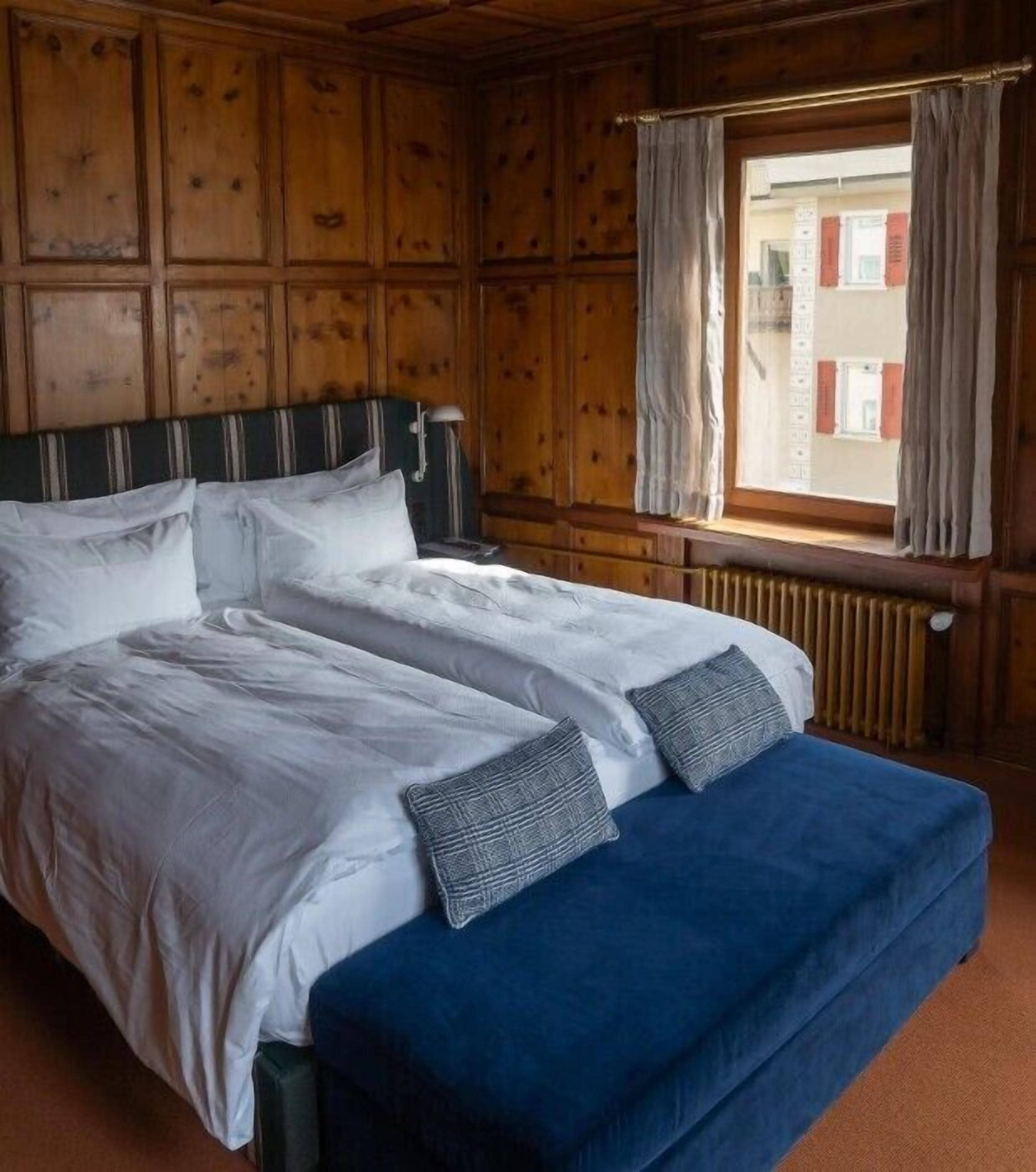 A double bed with white linens and two pillows, a blue bench, and a window with curtains.