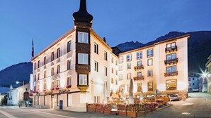 A hotel with a prominent corner tower, multiple balconies, and a well-lit entrance area with outdoor seating.