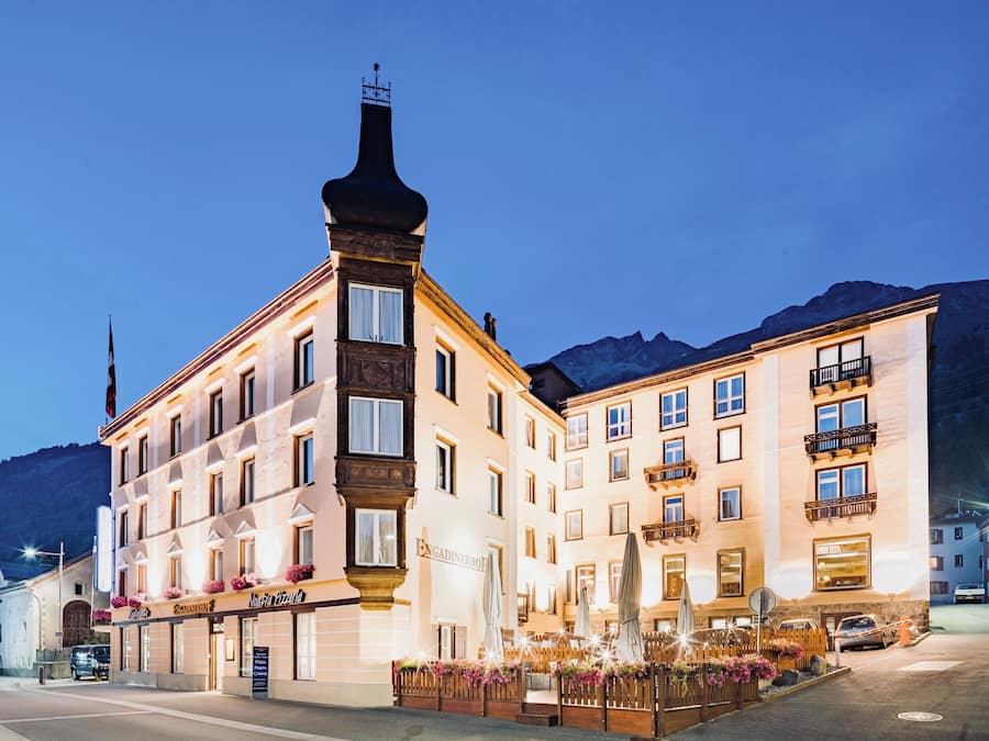 Ein Hotel mit einem markanten Eckturm, mehreren Balkonen und einem gut beleuchteten Eingangsbereich mit Außenbestuhlung.
