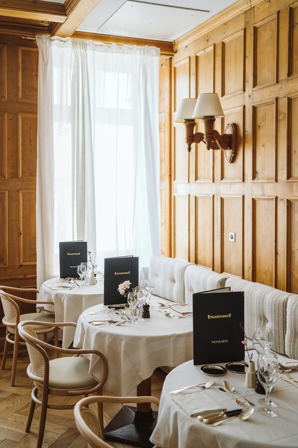 Un comedor con paredes de paneles de madera, cortinas blancas y mesas dispuestas con vajilla y cristalería fina.