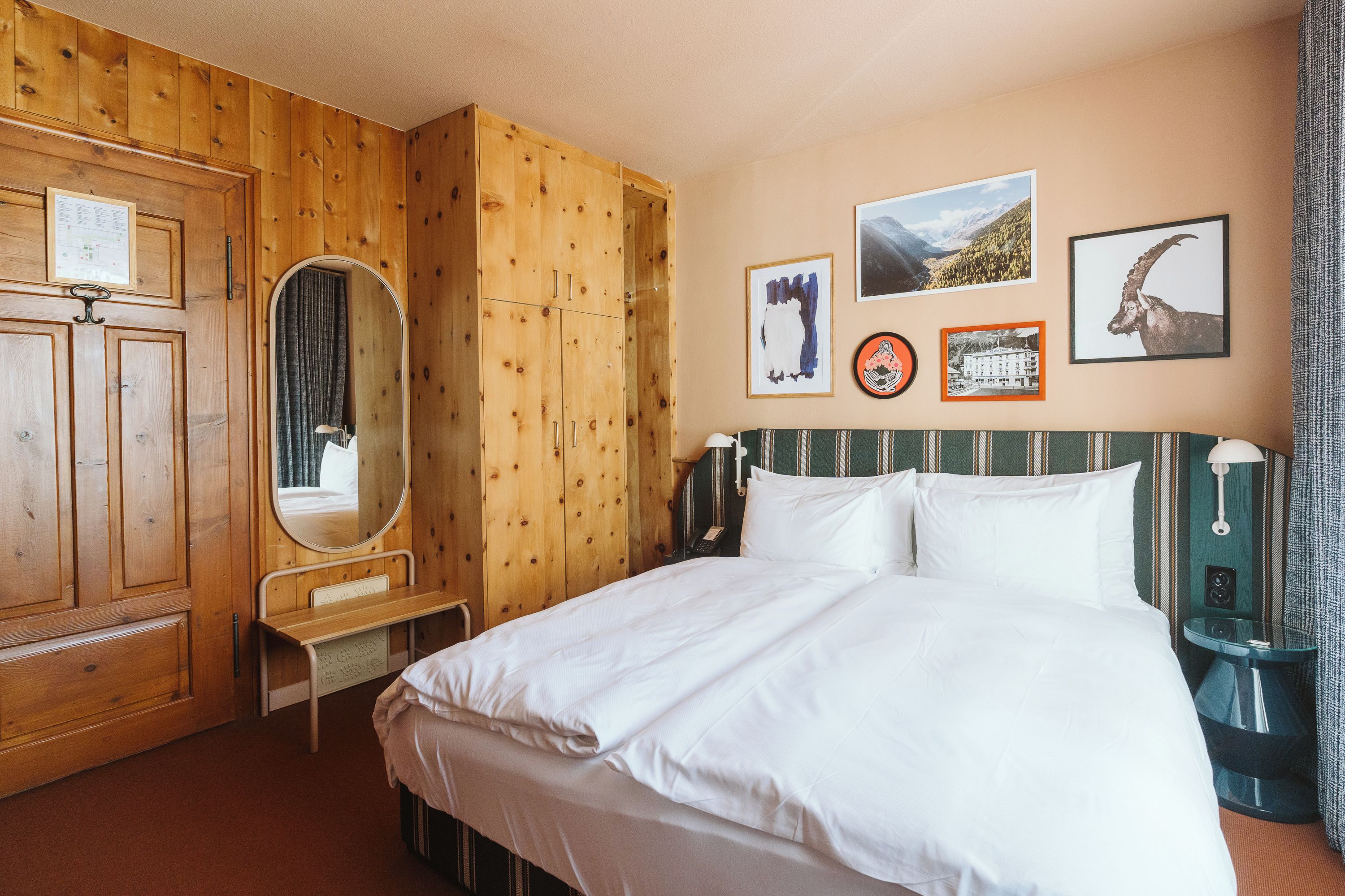 A hotel room with a wooden wardrobe, a bed with white bedding, a bedside table with a lamp, and a wall with framed pictures.