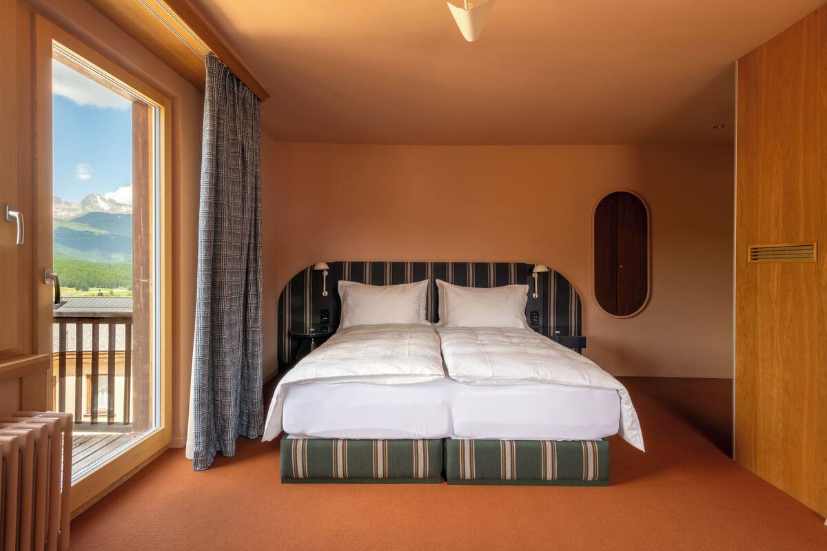 Een tweepersoonsbed met een gestreept hoofdeinde, uitzicht op bergen door een groot raam en een balkon met houten hekwerk.