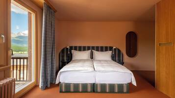 A double bed with a striped headboard, a view of mountains through a large window, and a balcony with a wooden railing.