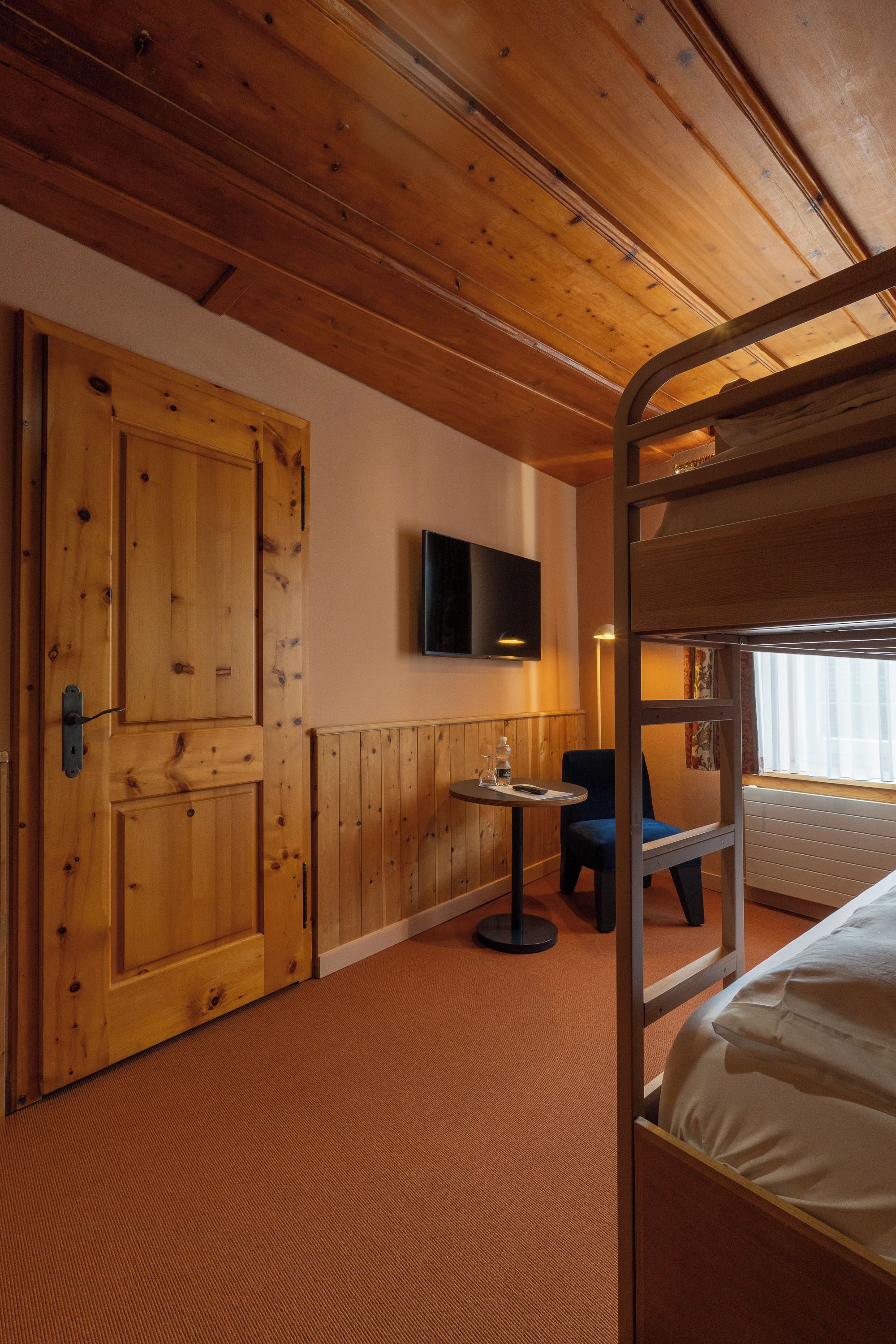 Habitación de hotel con literas, un televisor montado en la pared, una mesita con una silla y una puerta de madera.