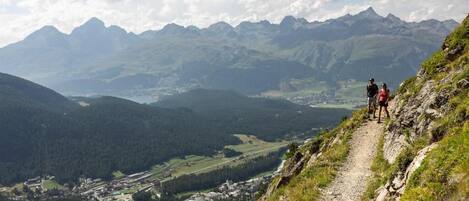 A mountain trail with hikers, overlooking a valley with buildings and a lake.