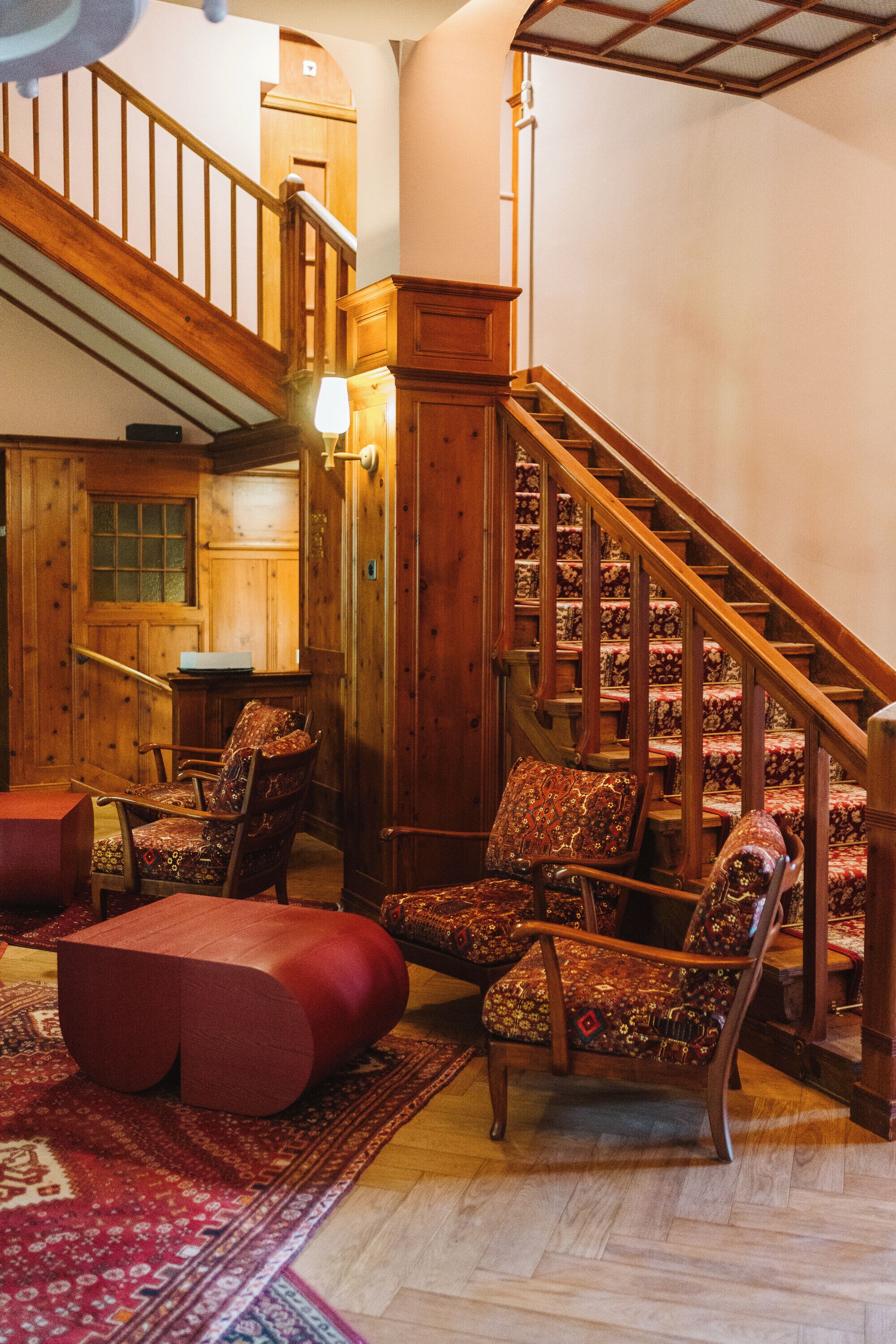 A wooden staircase with a patterned carpet, a wooden chair, and a red ottoman in a room with wooden walls and a wooden door.