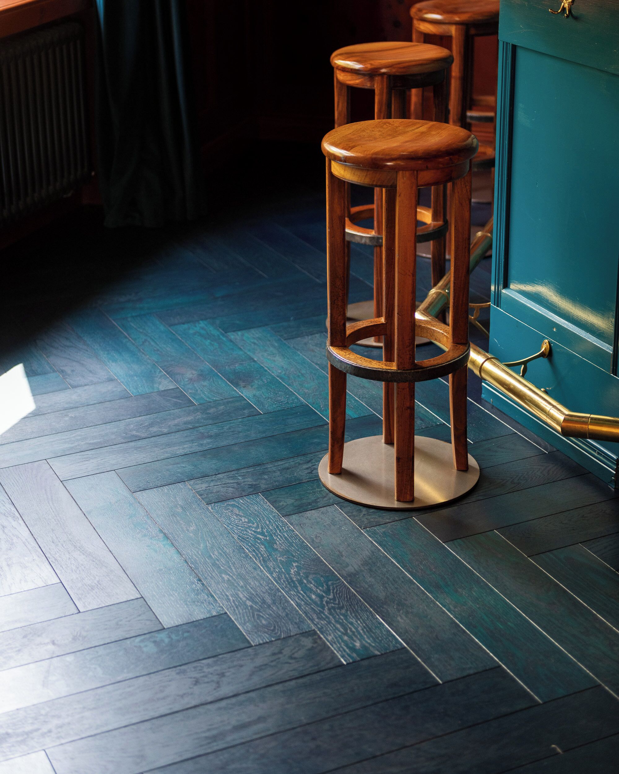 A wooden bar stool with a brass base, a teal-colored cabinet with a brass handle, and a herringbone-patterned wooden floor.