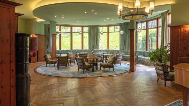 A spacious room with a large round table surrounded by chairs, large windows with curtains, and a wooden floor. - Boutique Hotel Engadinerhof – A Home for Generation (Pontresina)