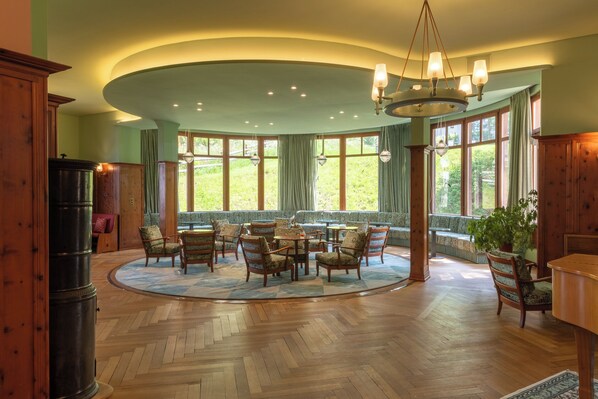 A spacious room with a large round table surrounded by chairs, large windows with curtains, and a wooden floor.