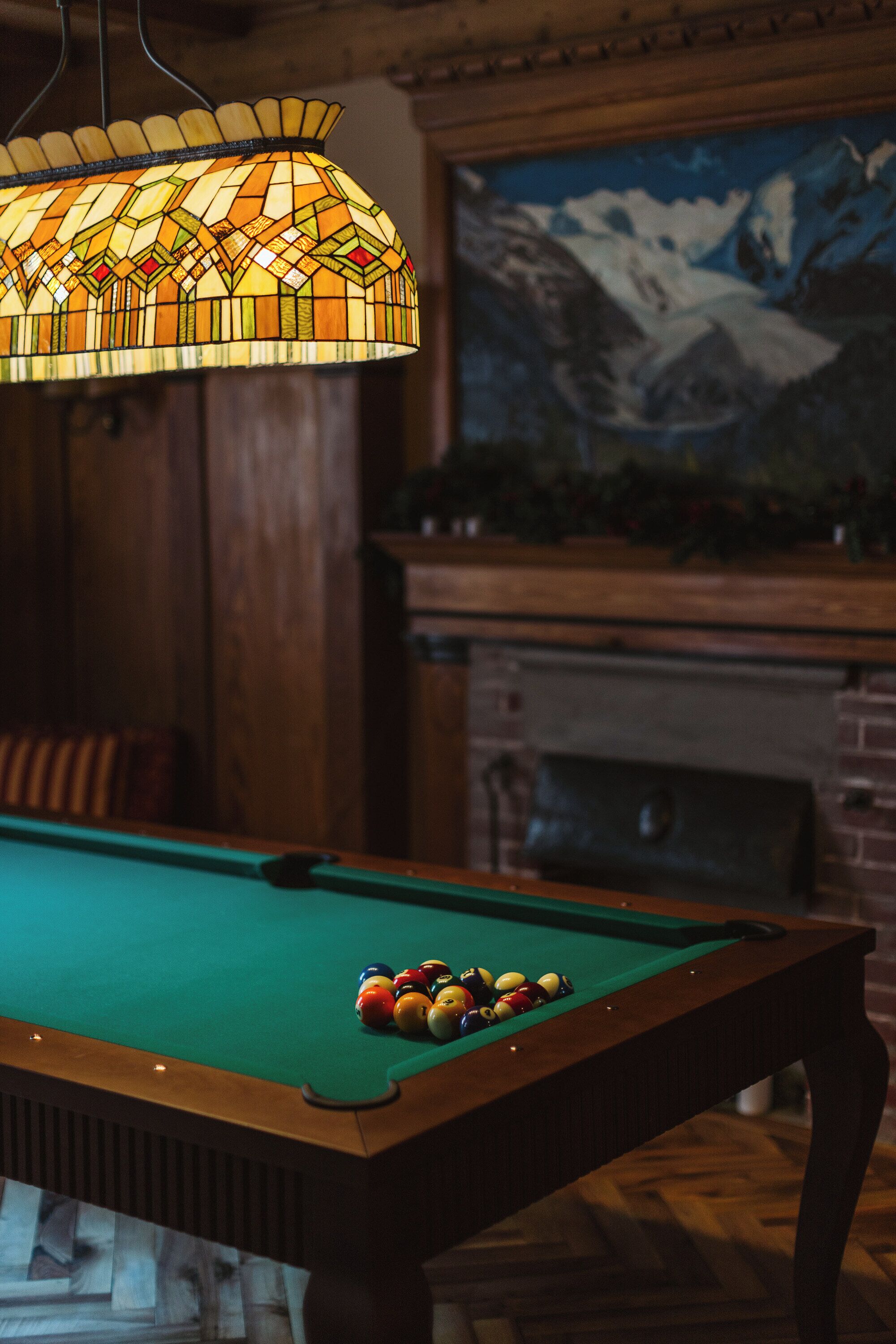 A pool table with balls, a stained glass lamp, and a fireplace with a painting above it.