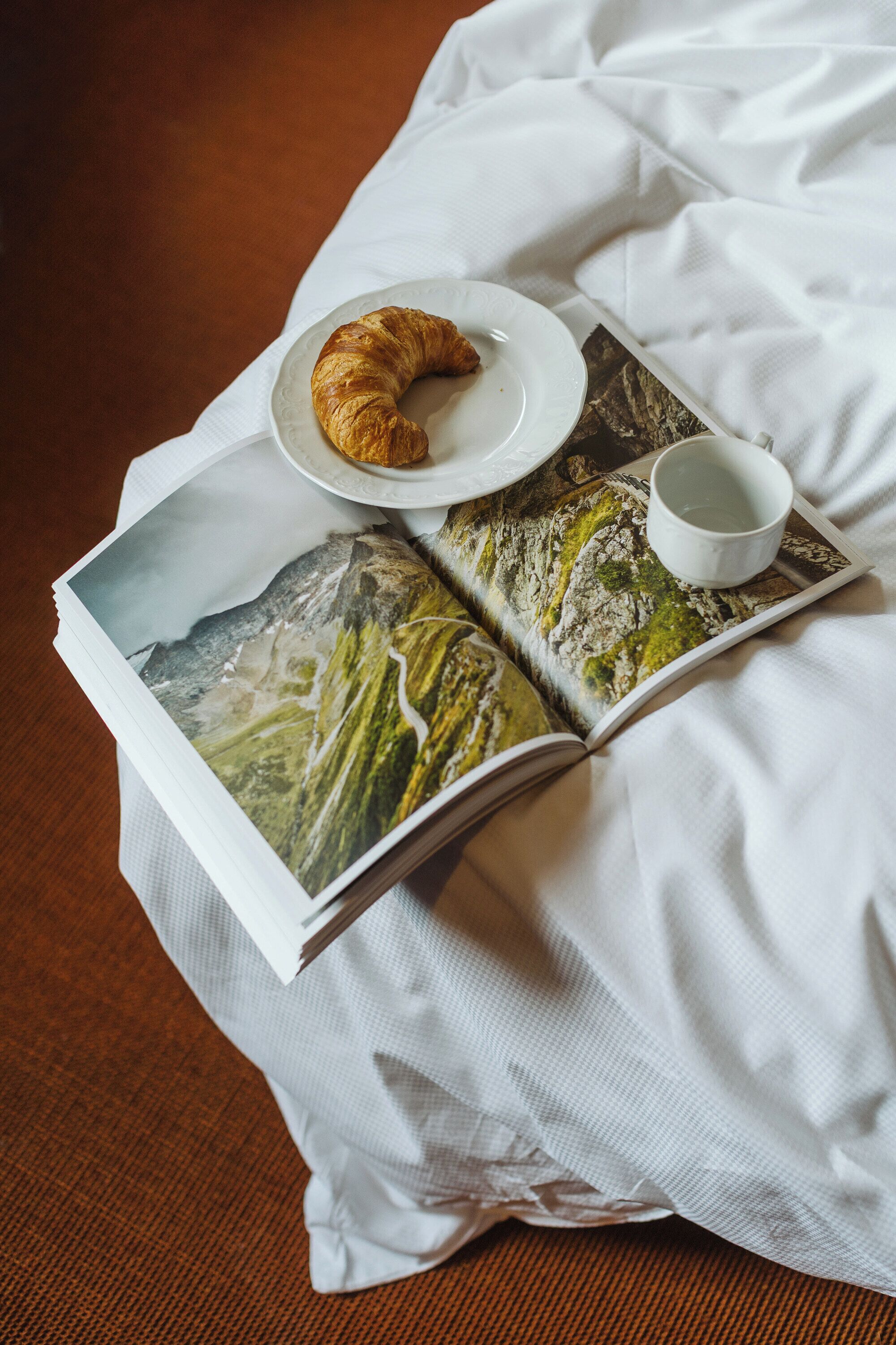 Eine Zeitschrift mit Berglandschaft, einem Croissant und einer Tasse auf einem Bett.