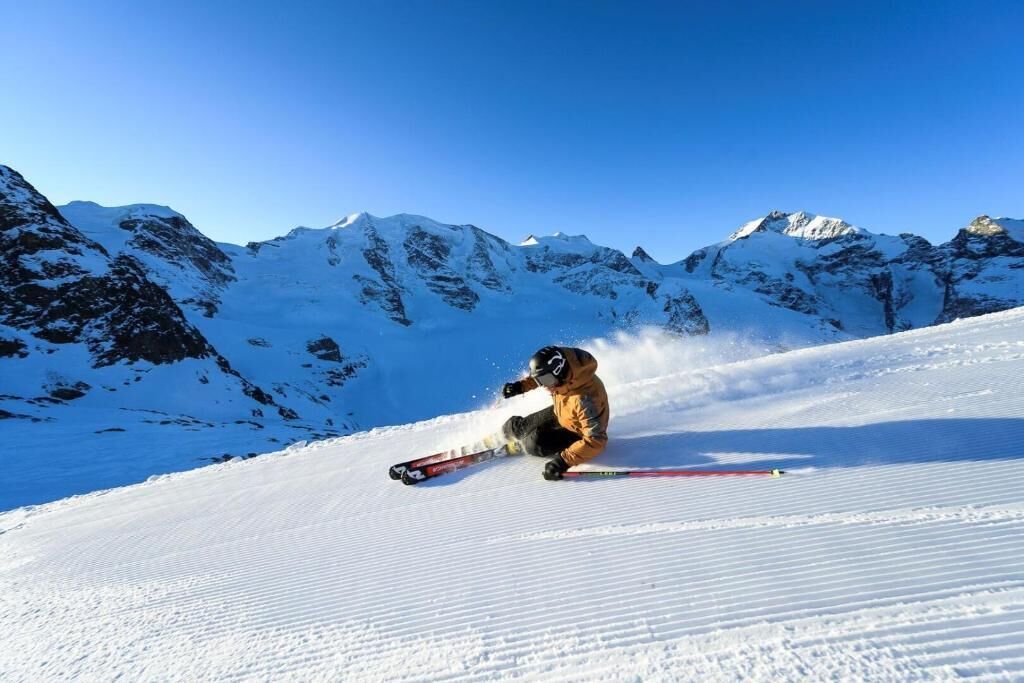 A skier in a crouched position on a snowy slope with a clear blue sky and snow-covered mountains in the background.