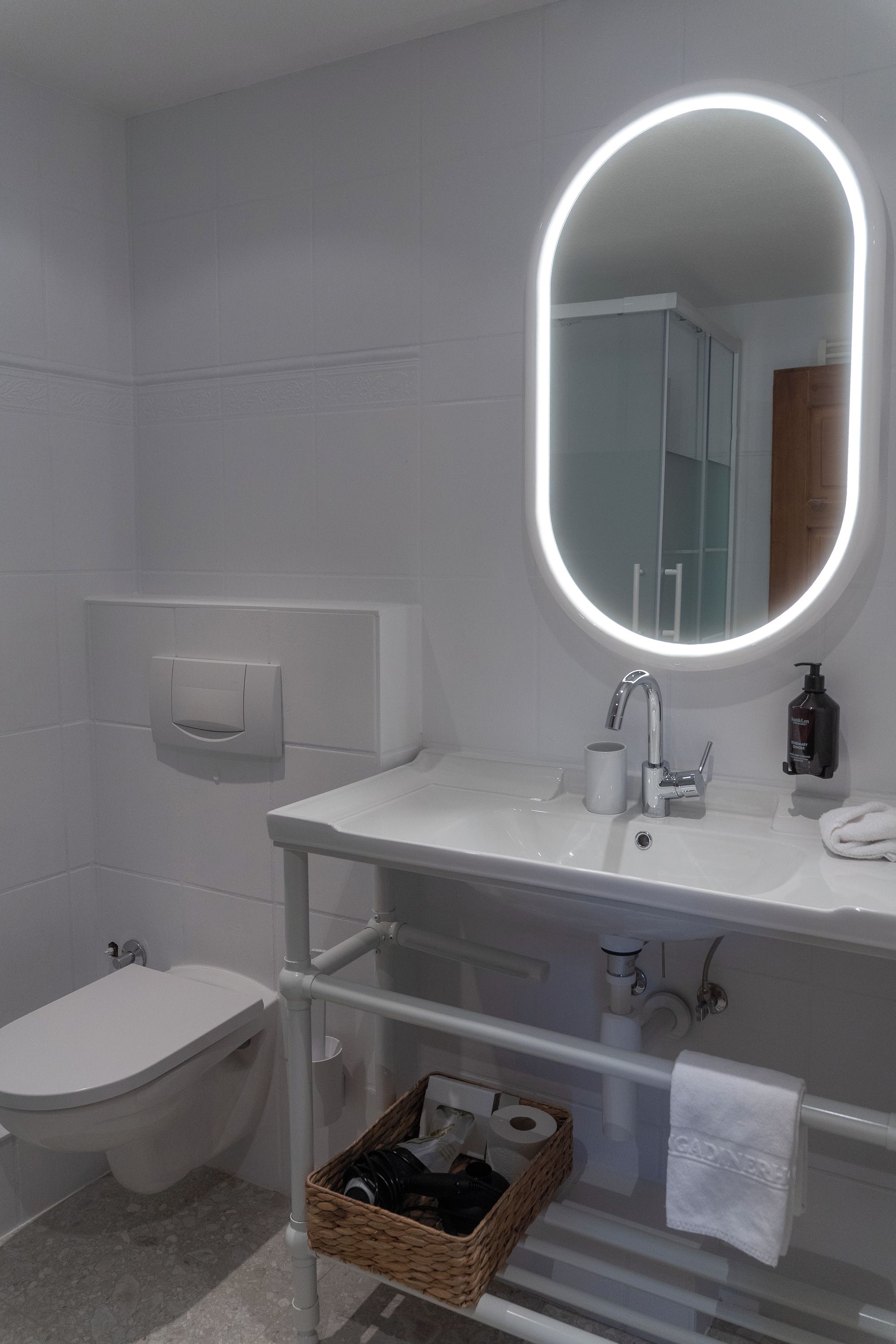 Uma casa de banho moderna com uma pia branca, um espelho iluminado, uma sanita e um cesto de produtos de higiene pessoal.