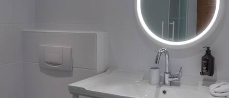 Uma casa de banho moderna com uma pia branca, um espelho iluminado, uma sanita e um cesto de produtos de higiene pessoal.