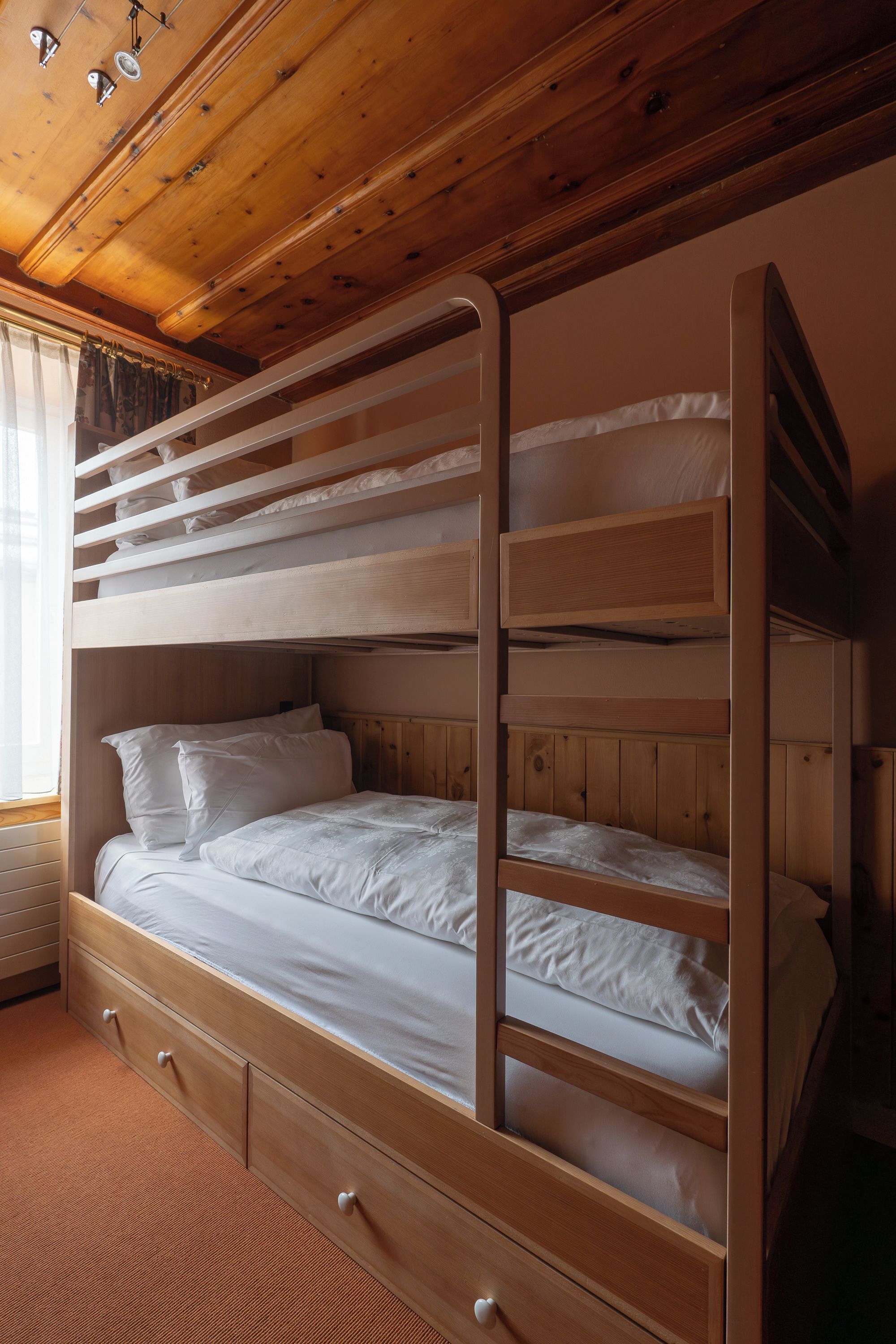 A bunk bed with a wooden frame, white bedding, and a window with curtains.
