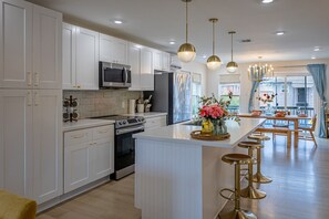 Una cocina moderna con armarios blancos, electrodomésticos de acero inoxidable, una isla central con taburetes, iluminación colgante y vistas al comedor.