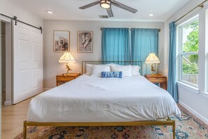 A bedroom with a bed, two bedside tables with lamps, a ceiling fan, a window with curtains, and a door.
