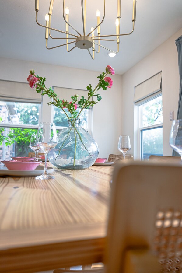 Una mesa de comedor con un florero de vidrio transparente con flores rosadas, una copa de vino y un bol rosado.