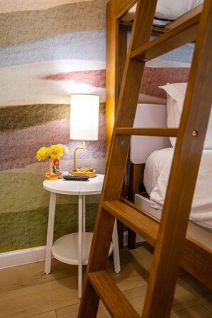 A bunk bed with a wooden ladder, a small white table with a lamp and a vase of flowers, and a colorful striped wall. - The Axelrod | Sleeps 16 (Houston)