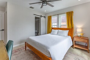 Un dormitorio con cama, mesita de noche, silla, ventilador de techo y ventana con cortinas.