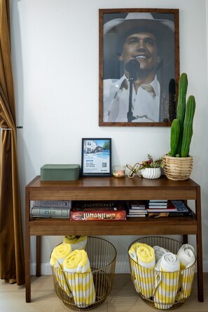 A wooden console table with a framed picture, a small potted plant, and a framed advertisement. Below, there are books and a basket with yellow towels. - The Axelrod | Sleeps 16 (Houston)