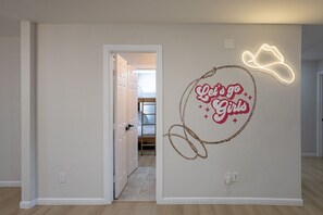 A room with a bunk bed, a neon sign on the wall, and a door leading to another room. - The Axelrod | Sleeps 16 (Houston)