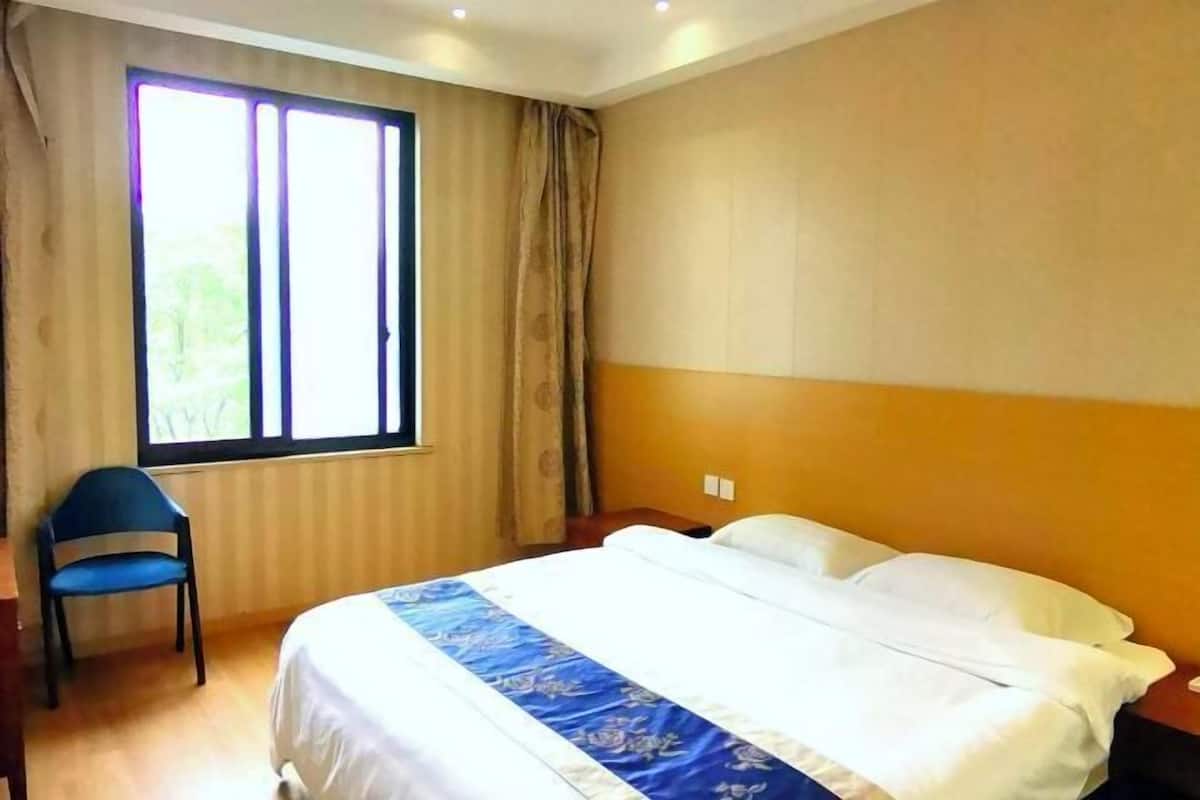 A hotel room with a bed, a chair, a window, and a wall with a light switch.