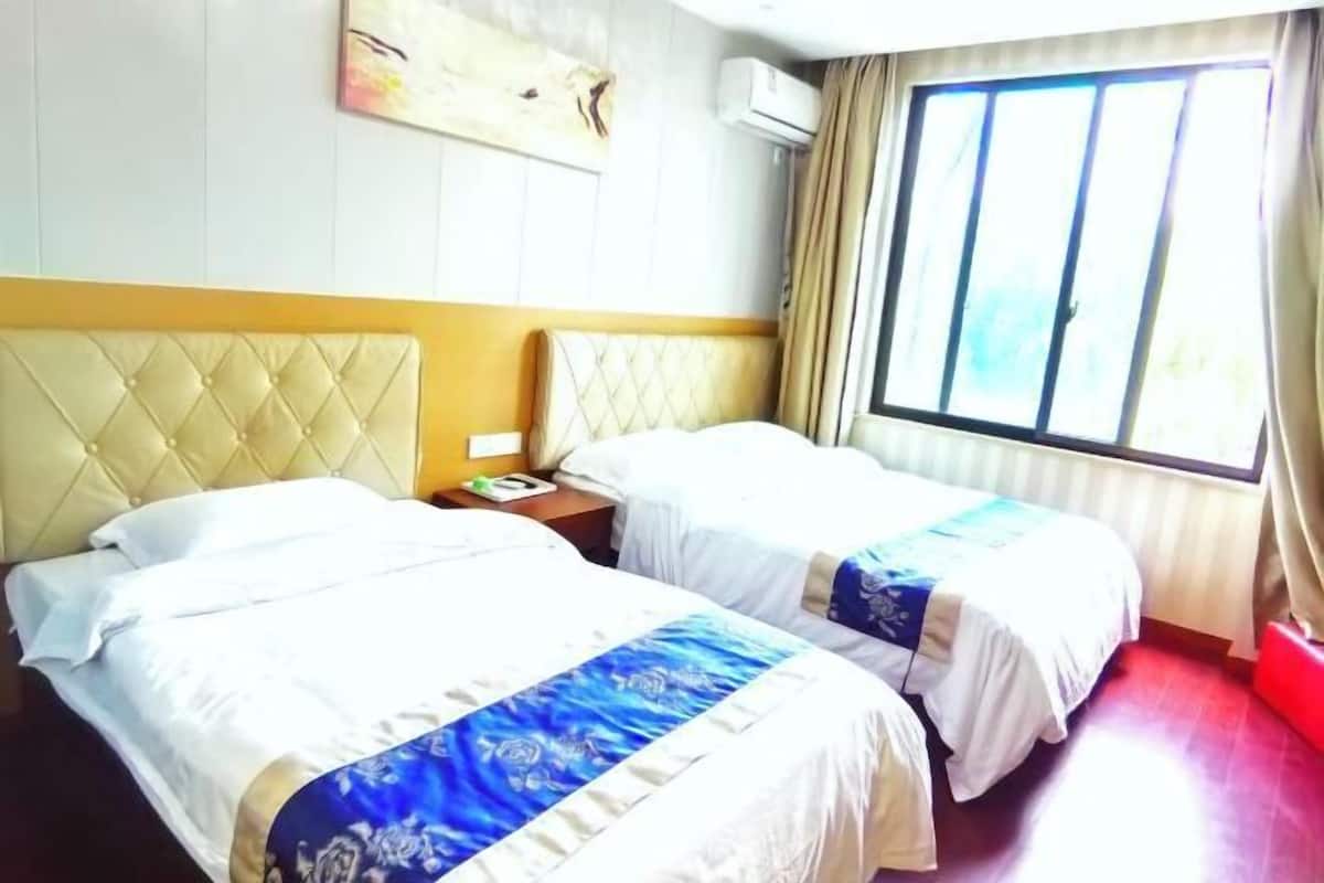 A hotel room with two beds, a headboard, a bedside table, a window with curtains, and an air conditioning unit.