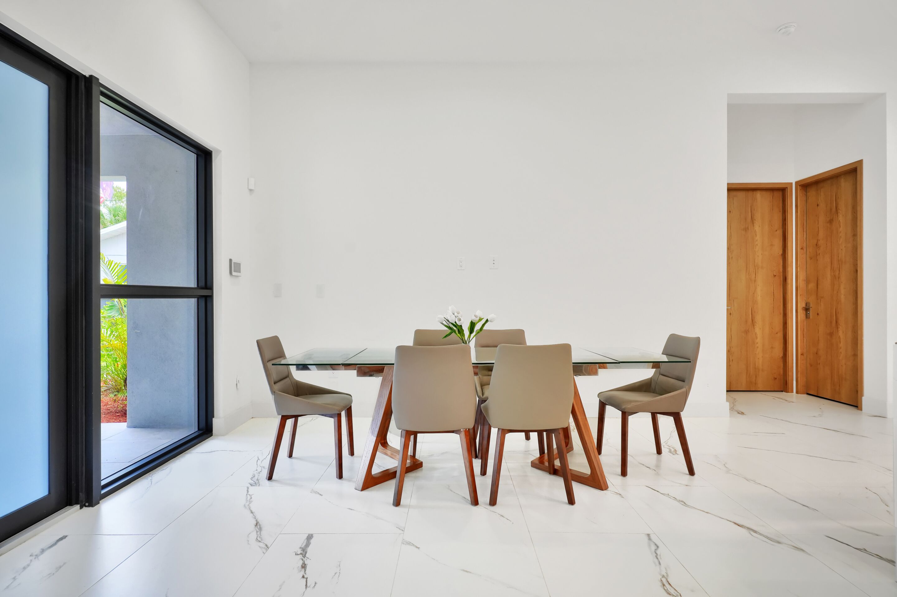 Un comedor moderno con una mesa de vidrio y seis sillas, ventanas amplias y una puerta de madera.