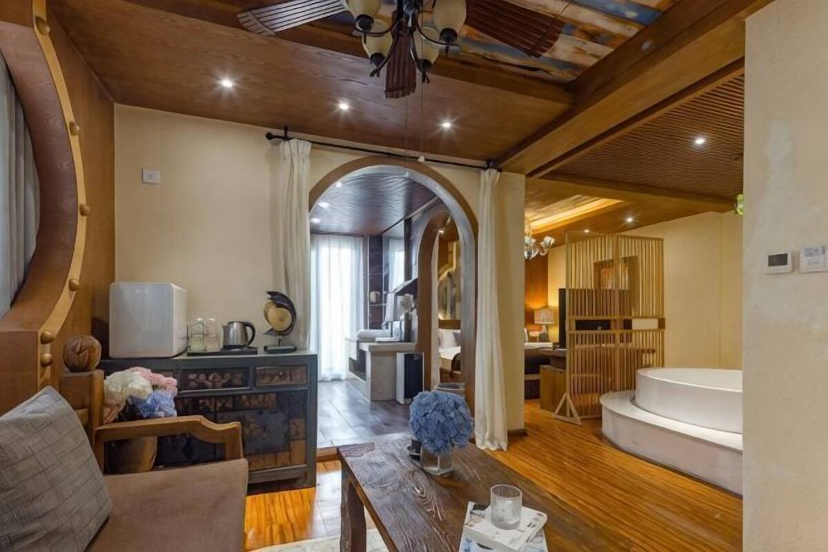 A spacious room with a wooden ceiling, a seating area with a sofa and a table, a large window with curtains, and a built-in bathtub.
