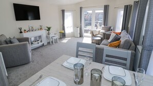 A modern living room with a dining area, grey sofas, a flat-screen TV, and a balcony with a view. - Casa De Rhu (Evesham)