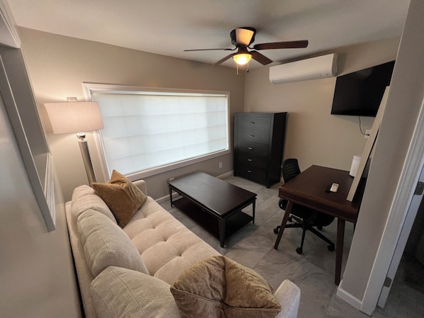 A compact living space with a sofa, a small table, a desk with a chair, a ceiling fan, and a TV mounted on the wall.