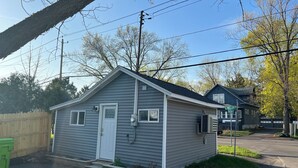 A small, single-story house with a gray exterior and a white door. It has a small front porch and a garage attached to the right. - Charming Tiny House minutes from Syracuse! (Syracuse)