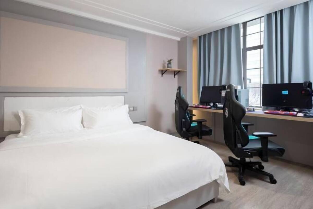 Een hotelkamer met een bed, twee bureaus met computers, een raam met gordijnen en een klein plankje met een plant.