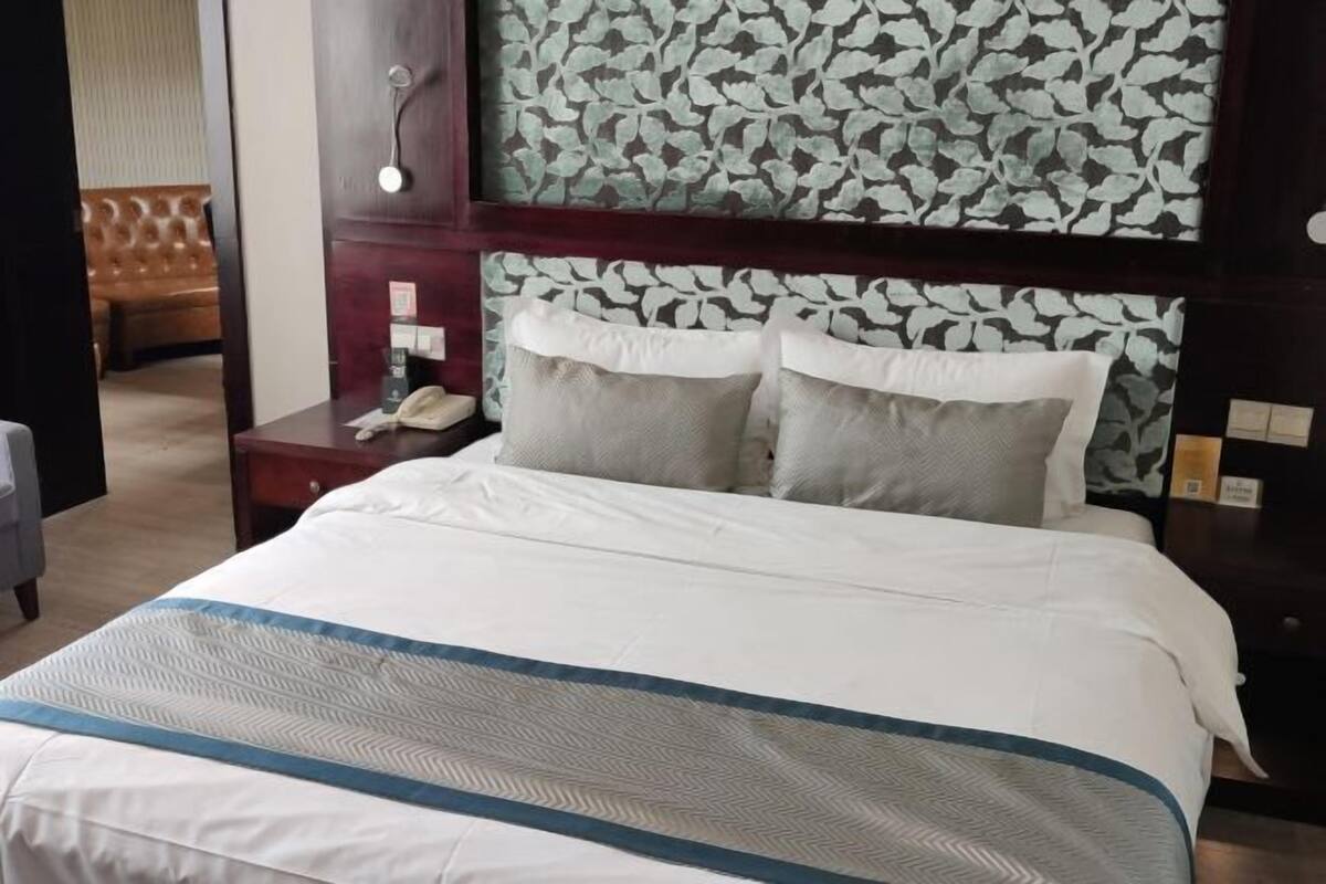 Een net opgemaakt bed met een wit-blauwe spreideken, een houten hoofdbord met een bladmotief en een nachtkastje met een telefoon en een kleine lamp.