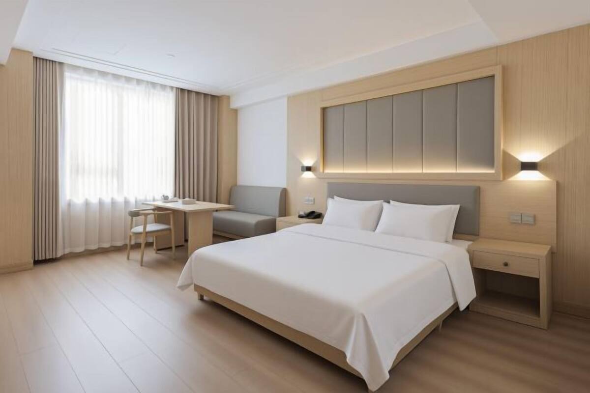 Een hotelkamer met een bed, bureau, stoel, nachtkastje en een groot raam met gordijnen.