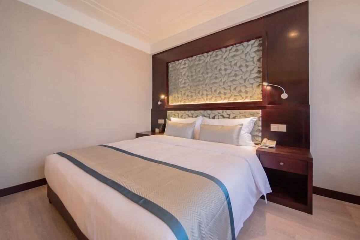 Een opgemaakt bed met een hoofdbord met patroon, een nachtkastje met een lamp en een wandlamp.